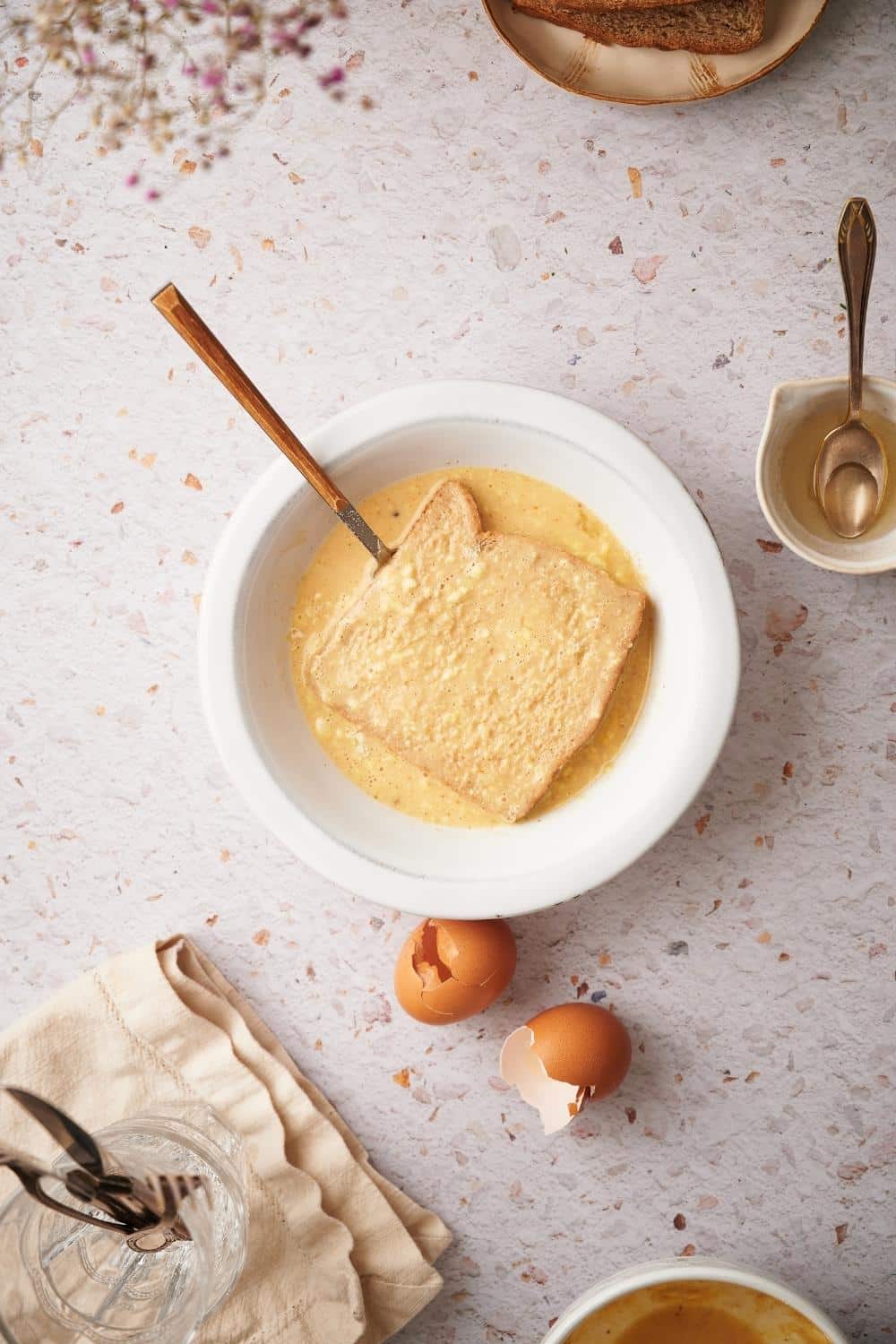 A slice of bread being soaked in french toast mixture in a bowl. Surrounding the bowl is a small syrup pitcher with a spoon and two cracked egg shells.