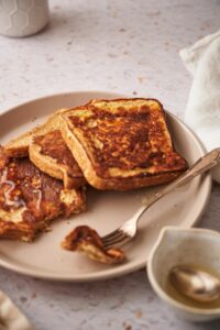 Close up of french toasts fanned out on a plate. The french toasts are drizzled with syrup and a fork with a piece of french toast is also on the plate.