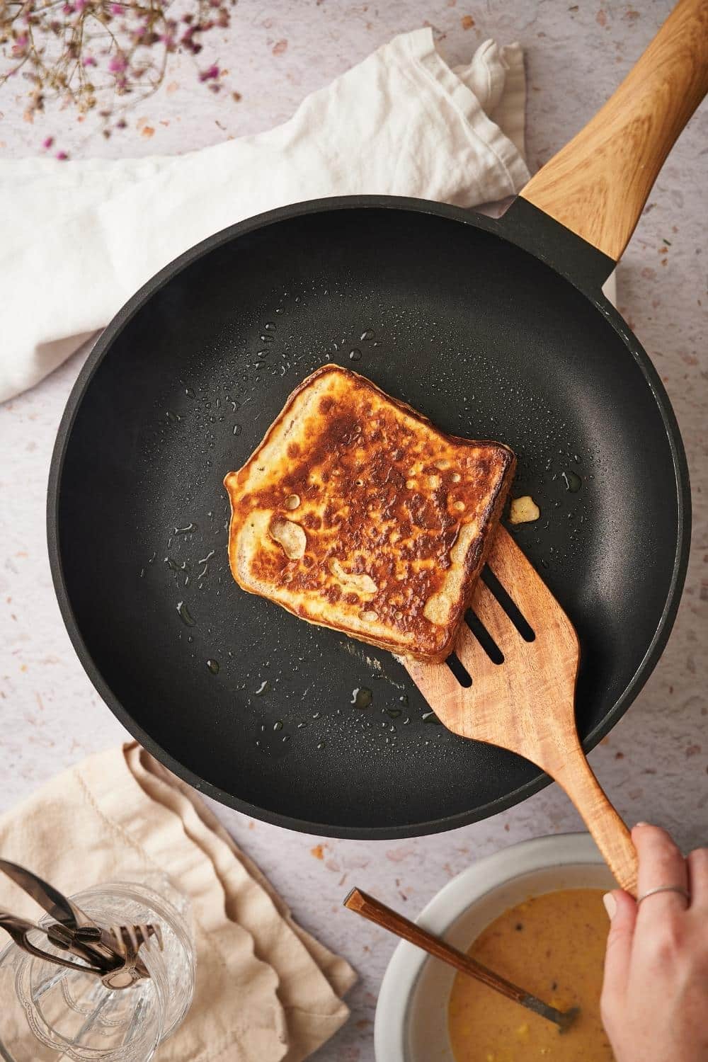 A cooked slice of french toast on a black pan with a wooden handle. A wooden spatula is being used to lift the french toast.