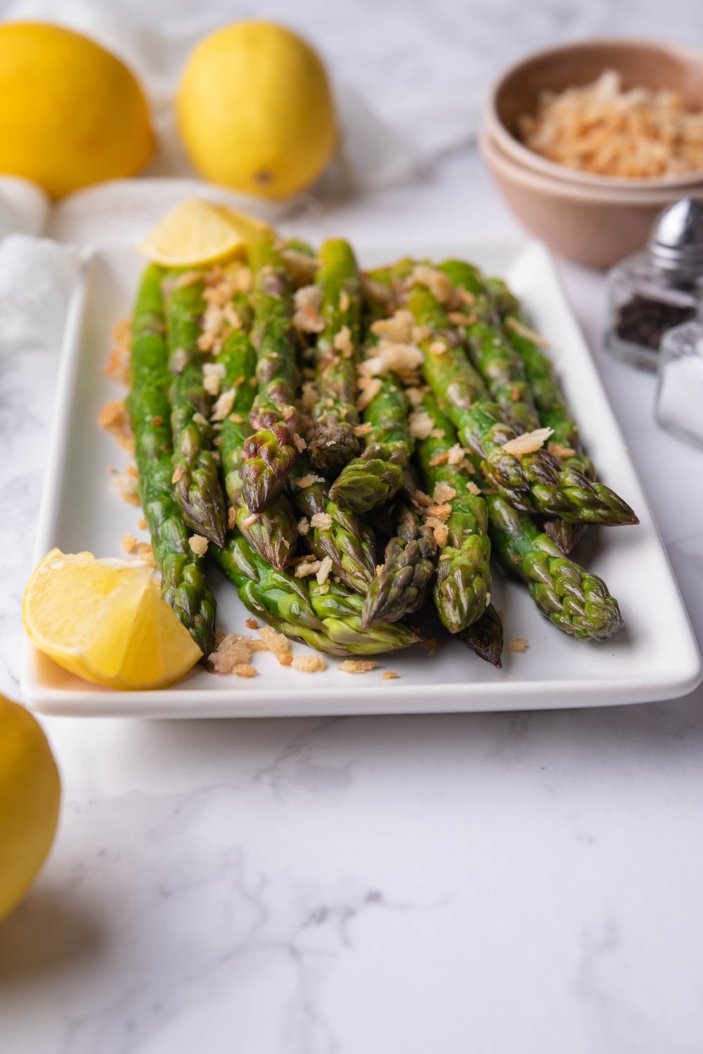 Sauteed asparagus spears on a white rectangle plate. The asparagus is garnished with panko breadcrumbs and served with lemon wedges. Behind it are whole lemon wedges on a tea towel, a small bowl of breadcrumbs, and salt and pepper shakers.