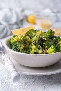 Close up of sauteed broccoli with red pepper flakes and grated parmesan in a white bowl garnished with a lemon wedge. The bowl is on top of a white plate next to a fork with a white handle. Behind it are more lemon wedges and a tea towel.