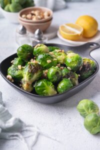 Sauteed brussels sprouts garnished with chopped hazlnuts in a small square cast iron skillet. Behind it is a bowl of fresh raw brussel sprouts, a bowl of hazelnuts, salt and pepper shakers, and a plate of lemon with half a lemon wedge.