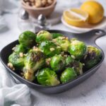Sauteed brussels sprouts garnished with chopped hazlnuts in a small square cast iron skillet. Behind it is a bowl of whole hazelnuts, salt and pepper shakers, and a plate of lemon with half a lemon wedge.