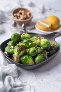 Sauteed brussels sprouts garnished with chopped hazlnuts in a small square cast iron skillet. Behind it is a bowl of whole hazelnuts, salt and pepper shakers, and a plate of lemon with half a lemon wedge.