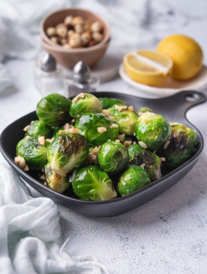 Sauteed brussels sprouts garnished with chopped hazlnuts in a small square cast iron skillet. Behind it is a bowl of whole hazelnuts, salt and pepper shakers, and a plate of lemon with half a lemon wedge.