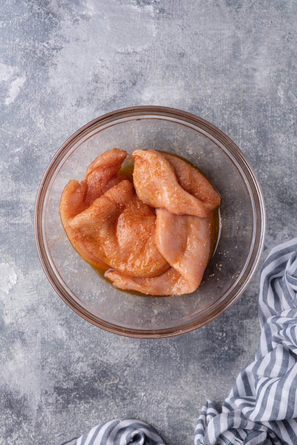 Raw sliced chicken breast coated with olive oil and seasonings in a glass bowl. A striped tea towel sits to the side.