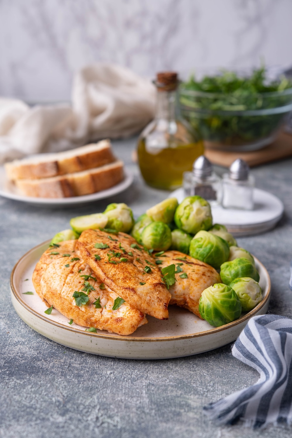 Sauteed chicken breasts garnished with herbs on a plate with steamed brussel sprouts. On the same table is a striped tea towel, a plate of sliced bread, a bottle of olive oil, salt and pepper shakers, and a glass bowl filled with greens.