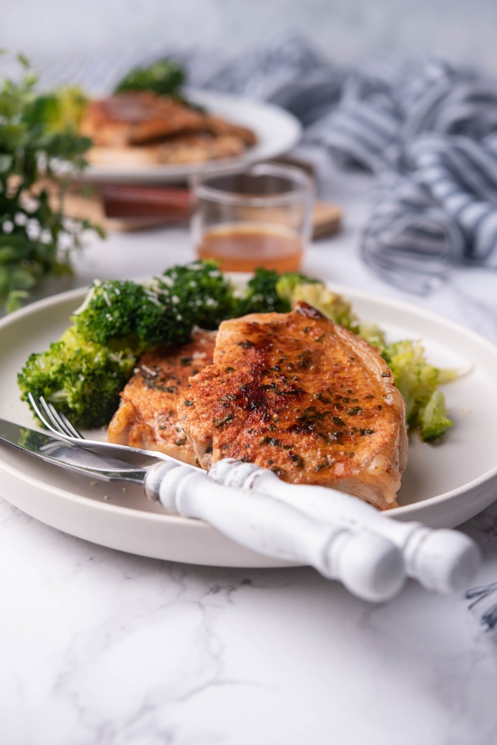 Thick sauteed pork chops with broccoli on a white plate with a fork and knife. Another plate of pork chops and broccoli can be seen in the background next to a glass sauce pitcher of pan sauce.