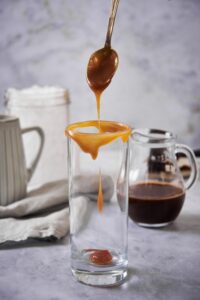 A metal spoon drizzling caramel sauce on the rim and the bottom of an empty tall glass. Behind it is a pitcher of cold brew, a pitcher of milk, and a large mason jar of ice.