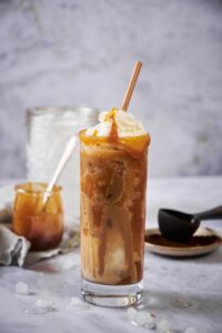 A tall glass of iced skinny caramel macchiato topped with whipped cream and a brown straw. Behind the glass is a small plate of ground coffee, a jar of ice, and a small jar of caramel.