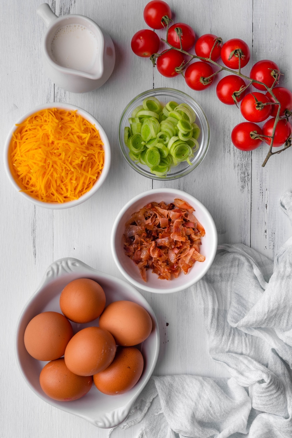 A bowl of six eggs, a bowl of crumbled bacon, a bowl of slices green onion, a vine of tomatoes, and a bowl of shredded cheddar cheese all on a white counter.