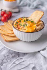 Healthy chicken dip in a bowl, garnished with parsley and topped with a cracker for dipping. The bowl is on a plate with more crackers. In the back are cherry tomatoes on a stem.