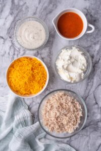 Glass bowls of cream cheese, ranch, and shredded chicken, and white bowls of hot sauce and cheddar cheese.