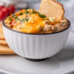 Healthy chicken dip in a bowl, garnished with parsley and topped with a cracker for dipping. The bowl is on a plate with more crackers.