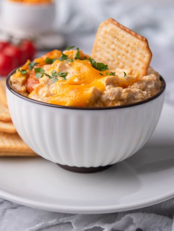 Healthy chicken dip in a bowl, garnished with parsley and topped with a cracker for dipping. The bowl is on a plate with more crackers.