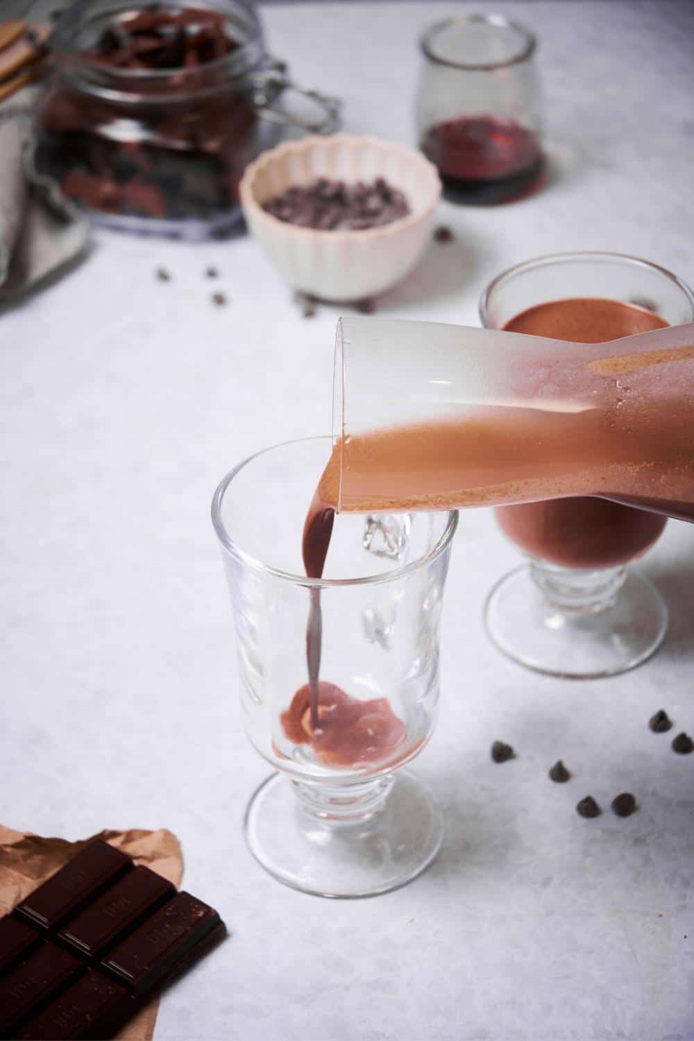 Hot chocolate being poured from a glass carafe into glass mugs. Scattered chocolate chips decorate the table, and surrounding the mugs is a bar of baking chocolate, a jar of chopped chocolate, a small bowl of chocolate chips, and a small jar of maple syrup.