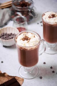 A closer look at a glass mug of hot chocolate topped with whipped cream and chocolate shavings. In the back is another glass of hot chocolate, a jar of chocolate, a small bowl of chocolate chips, and a small glass container of maple syrup. In front, part of a piece of baking chocolate can be seen.