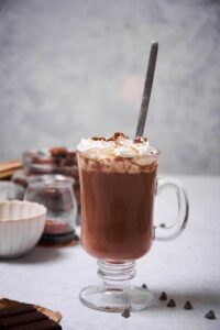 A glass mug of hot chocolate topped with whipped cream and chocolate shavings with a metal mixing spoon. On the table there are scattered chocolate chips. Surrounding the glass is a bar of baking chocolate, a jar of chopped chocolate, a bowl of chocolate chips, and a small jar of maple syrup.