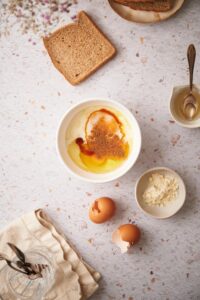 Unmixed french toast batter in a bowl, without the protein powder. Surrounding the bowl is a slice of whole wheat bread, a plate of bread, a small syrup pitcher with a spoon, a small bowl of protein powder, and two empty cracked egg shells.