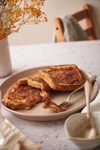 Three protein french toast slices fanned out on a plate. The french toasts are drizzled with syrup and a fork with a piece of french toast rests on the plate. In the foreground is a small empty syrup pitcher with a metal spoon.