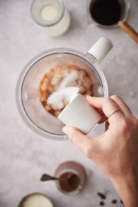 A hand pouring granulated keto sweetener into a blender of coffee and ice.