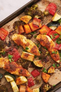 Three baked chicken tenderloins on a baking sheet lined with parchment paper surrounded by roasted broccoli, cucumber, and carrots.