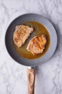 Seared pork chops over oil in a speckled grey pan with a wooden handle.