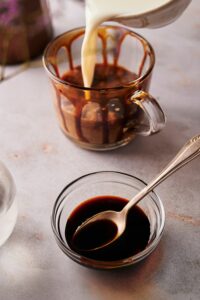 Milk being poured into a glass. In front of it is a bowl of caramel sauce with a spoon in it.