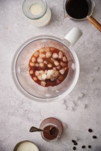 A blender with filled coffee and ice. Surrounding it is a jar of caramel sauce with a spoon, a carafe of almond milk, and a glass coffee pitcher filled with coffee.