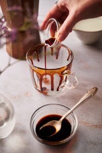 A hand pouring caramel sauce into a glass that is rimmed with the caramel sauce. A bowl of the caramel sauce is in front of it.