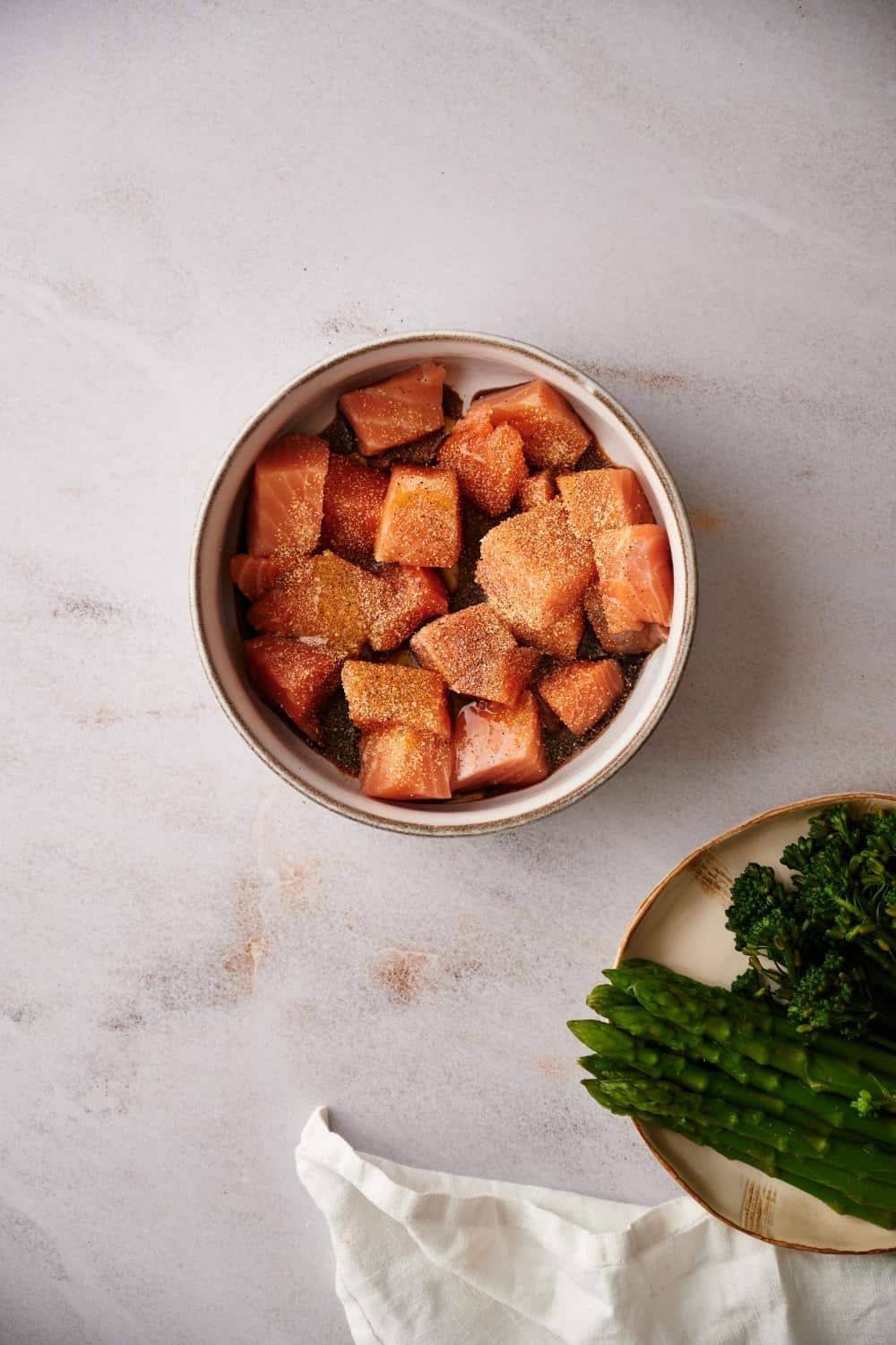 Raw seasoned salmon cubes in a small bowl. At the lower corner, part of a plate of asparagus and broccolini can be seen next to a tea towel.