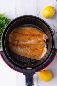 Raw seasoned tilapia fillets in an air fryer basket. Surrounding the basket are some whole lemons and a bundle of fresh parsley.