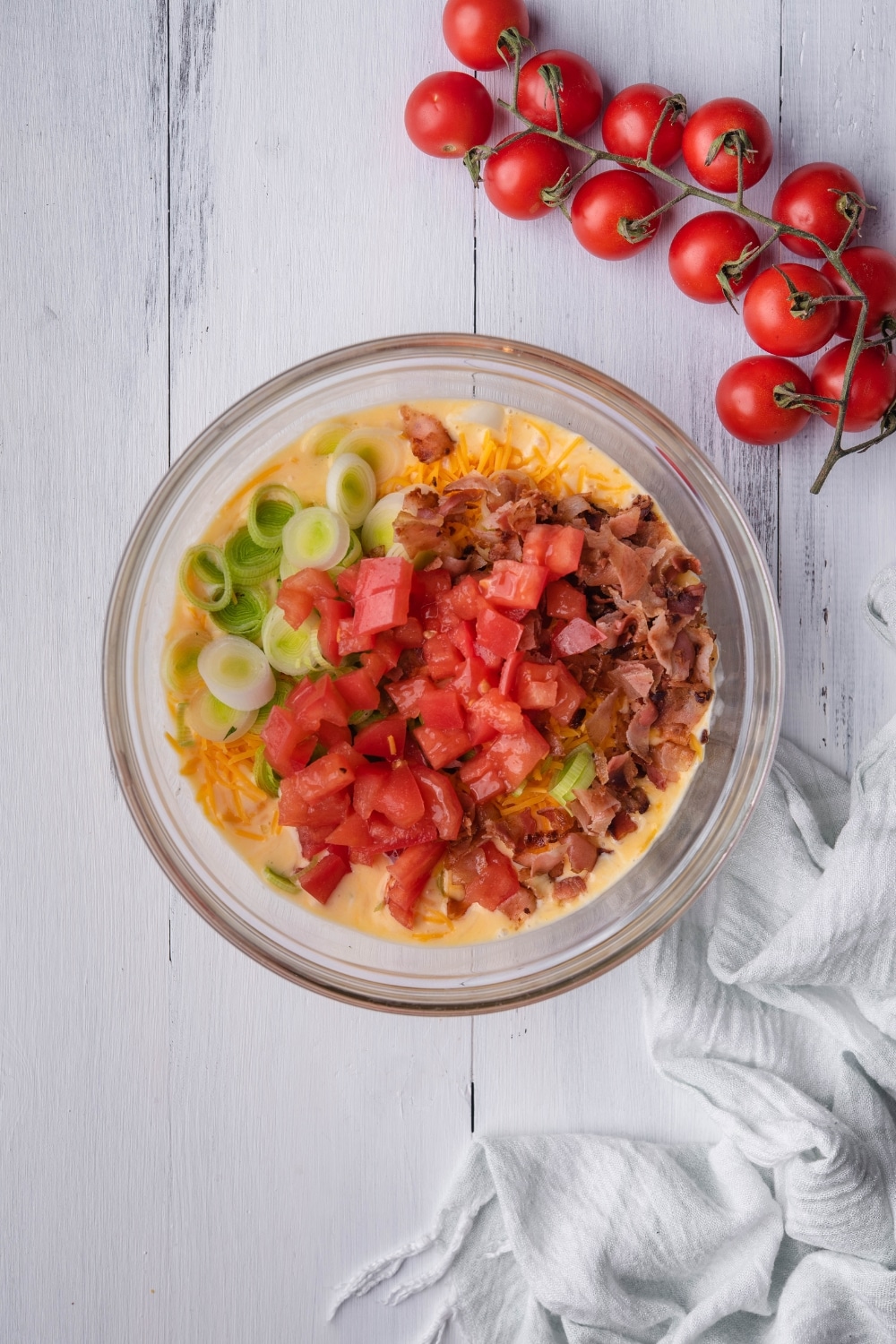 Diced tomatoes, green onion, bacon, and shredded cheese on top of whisked egg in a bowl.