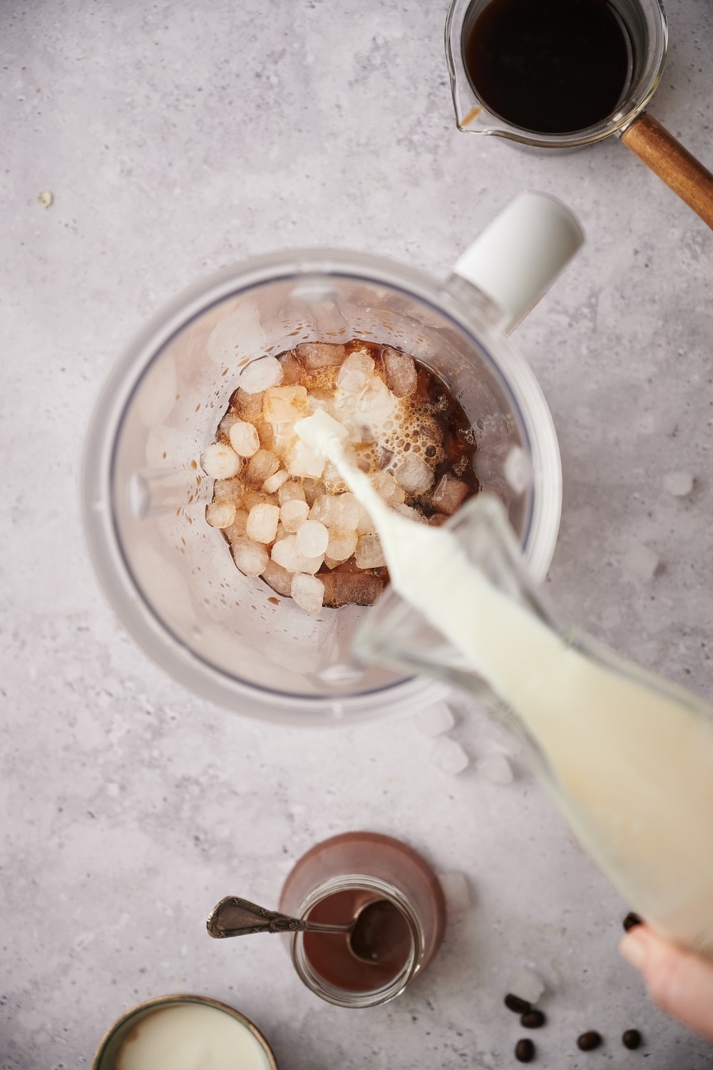 Almond milk being poured from a carafe into a blender of ice and coffee. Next to the blender is a glass coffee pitcher filled with coffee and a jar of keto caramel sauce.