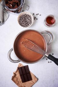 A metal whisk mixing hot chocolate in a saucepan. Surrounding the saucepan is a bar of baking chocolate, a small jar of chopped chocolate, a small bowl of chocolate chips, a small glass container of maple syrup, and a bar of baking chocolate.