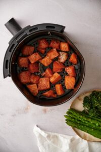 Raw seasoned salmon cubes in an air fryer basket. The salmon is in one even layer with a little spacing between each piece. Next to the basket is a plate of asparagus and broccolini and a white tea towel.