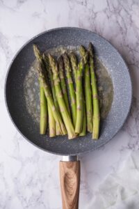 Raw asparagus sauteeing in butter and garlic in a grey speckled pan with a wooden handle. The pan is resting on a marble countertop.