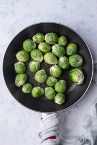 Raw brussels sprouts in a black pan. The handle of the pan is wrapped in a tea towel and the pan is resting on a grey countertop.