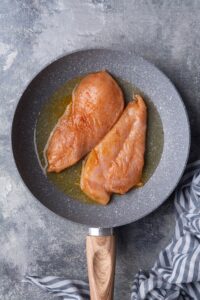 Two raw seasoned chicken breasts in a skillet with olive oil. The skillet is speckled dark grey with a wooden handle and it sits on a grey countertop next to a blue and white striped tea towel.