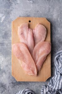 Two chicken breasts cut into 4 equally sized slices, fanned out on a wood cutting board. The board is on a grey countertop next to a blue and white striped tea towel.