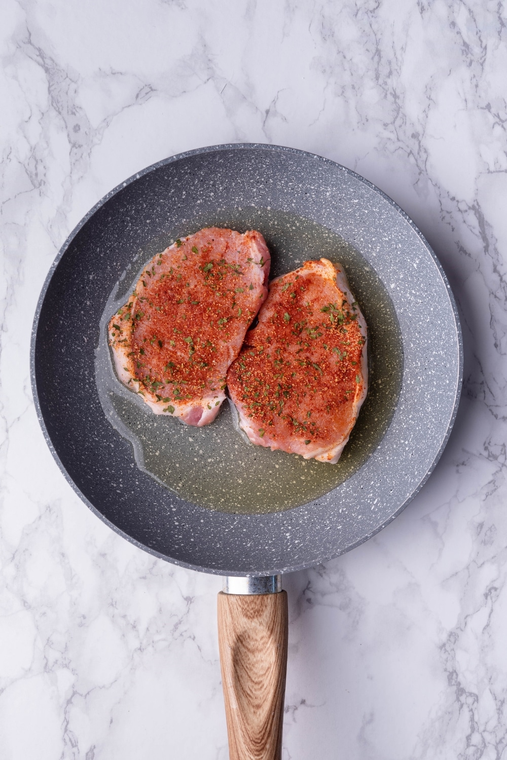 Uncooked seasoned pork chops over oil in a speckled grey pan with a wooden handle.