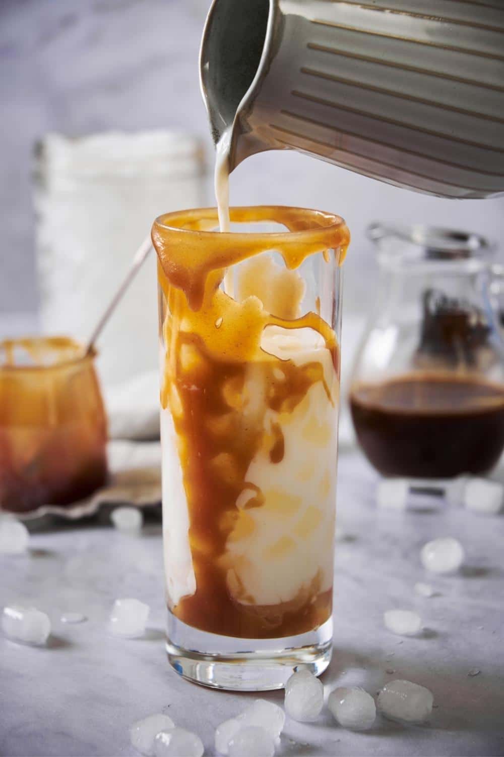 Almond milk being poured from a ceramic pitcher into a tall glass of ice and caramel syrup. Behind it is a pitcher of cold brew, a large mason jar of ice, and a small jar of caramel with a spoon in it.