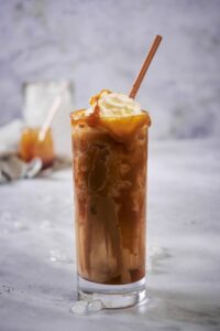 Mixed caramel macchiato in a tall glass. It's topped with whipped cream and caramel drizzle with a brown straw. Small pieces of ice are scattered across the counter and jar of ice and caramel can be seen at the back.