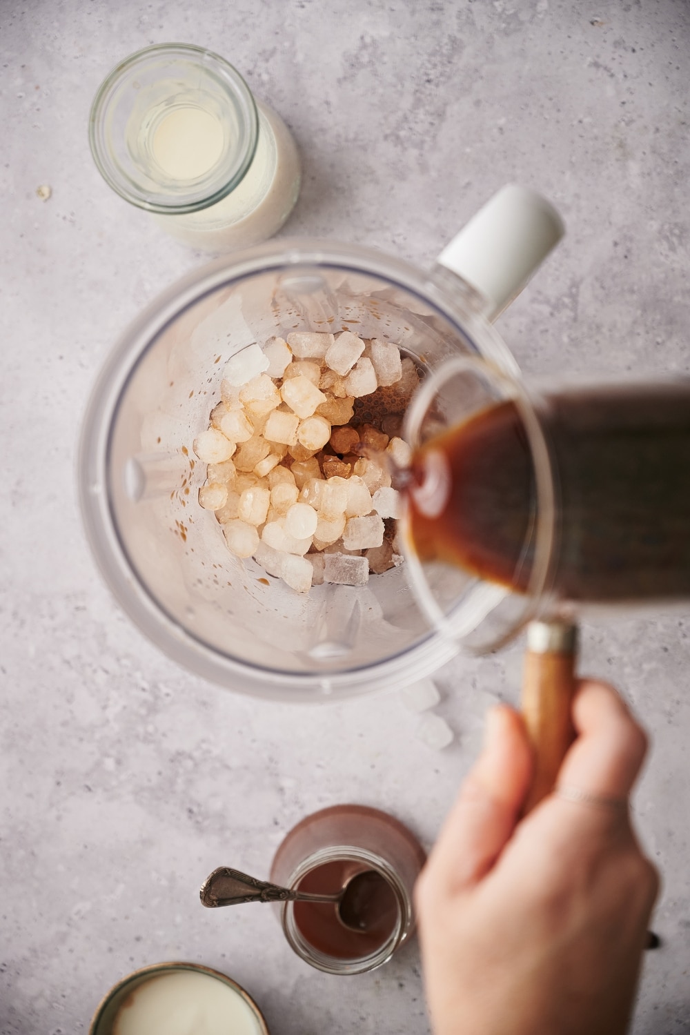 Coffee being poured from a glass coffee pitcher into a blender filled with ice. Next to the blender is a carafe of almond milk and a jar of keto caramel sauce.