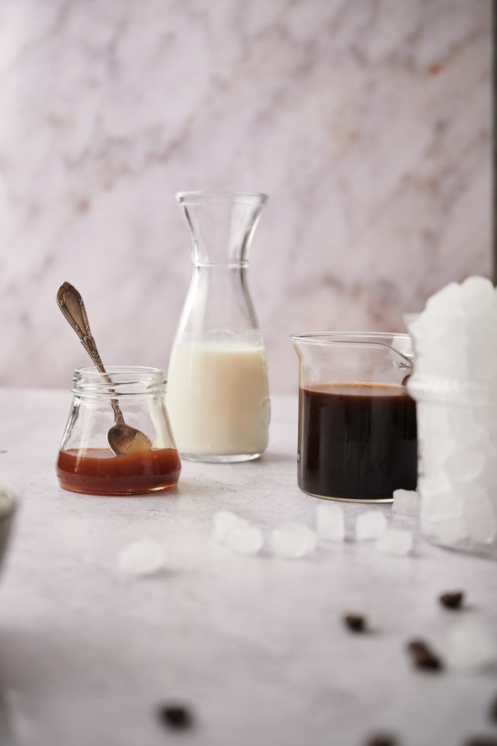 A carafe of almond milk, a small jar of keto caramel sauce with a spoon, a glass coffee pitcher filled with coffee, and a glass jar of nugget ice.