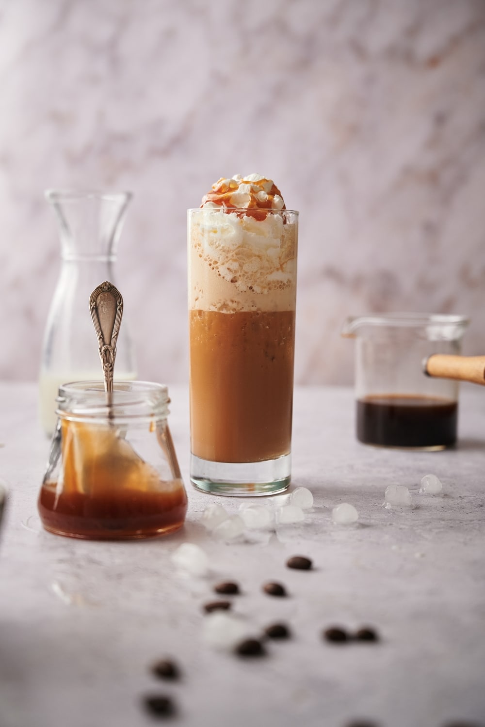 Keto frappuccino in a tall skinny glass topped with whipped cream and caramel sauce. Surrounding it is a jar of caramel sauce with a spoon, a half filled carafe of almond milk, and a glass coffee pitcher half filled with coffee.