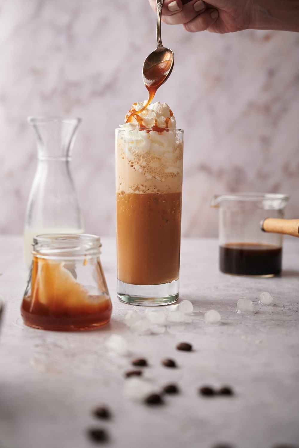 Keto caramel sauce being drizzled on a tall skinny glass of keto frappuccino with whipped cream. Surrounding it is a jar of caramel sauce, a half filled carafe of almond milk, and a glass coffee pitcher half filled with coffee.