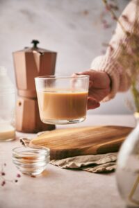 A hand picking up a glass mug of keto vanilla latte. Below it is an oval wooden board on top of folded tea towels. Surrounding the board are two small almost empty bowls, an almost empty bottle of almond milk, and a moka pot.