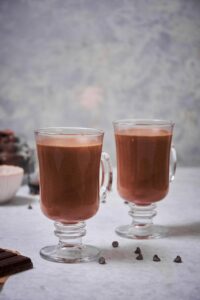 two glass mugs of hot chocolate. A few chocolate chips are sprinkled on the table and part of a bar of chocolate can be seen in the foreground.