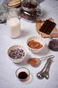 Hot chocolate ingredients. Clockwise from top: a jar of chopped baking chocolate, a bar of baking chocolate resting on a brown wrapper, a small jar of maple syrup, a small of cocoa powder, a measuring spoon filled with cocoa powder, smaller measuring spoons, a small glass bowl of vanilla extract, a small bowl of chocolate chips, and a glass bottle of milk.